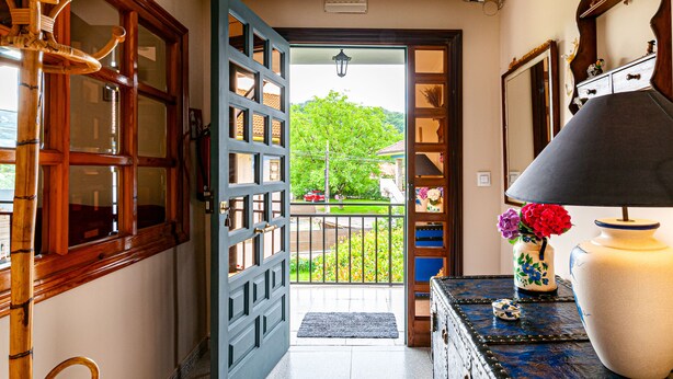 A hallway with a wooden door leading to a balcony with a view of a tree and a red car.