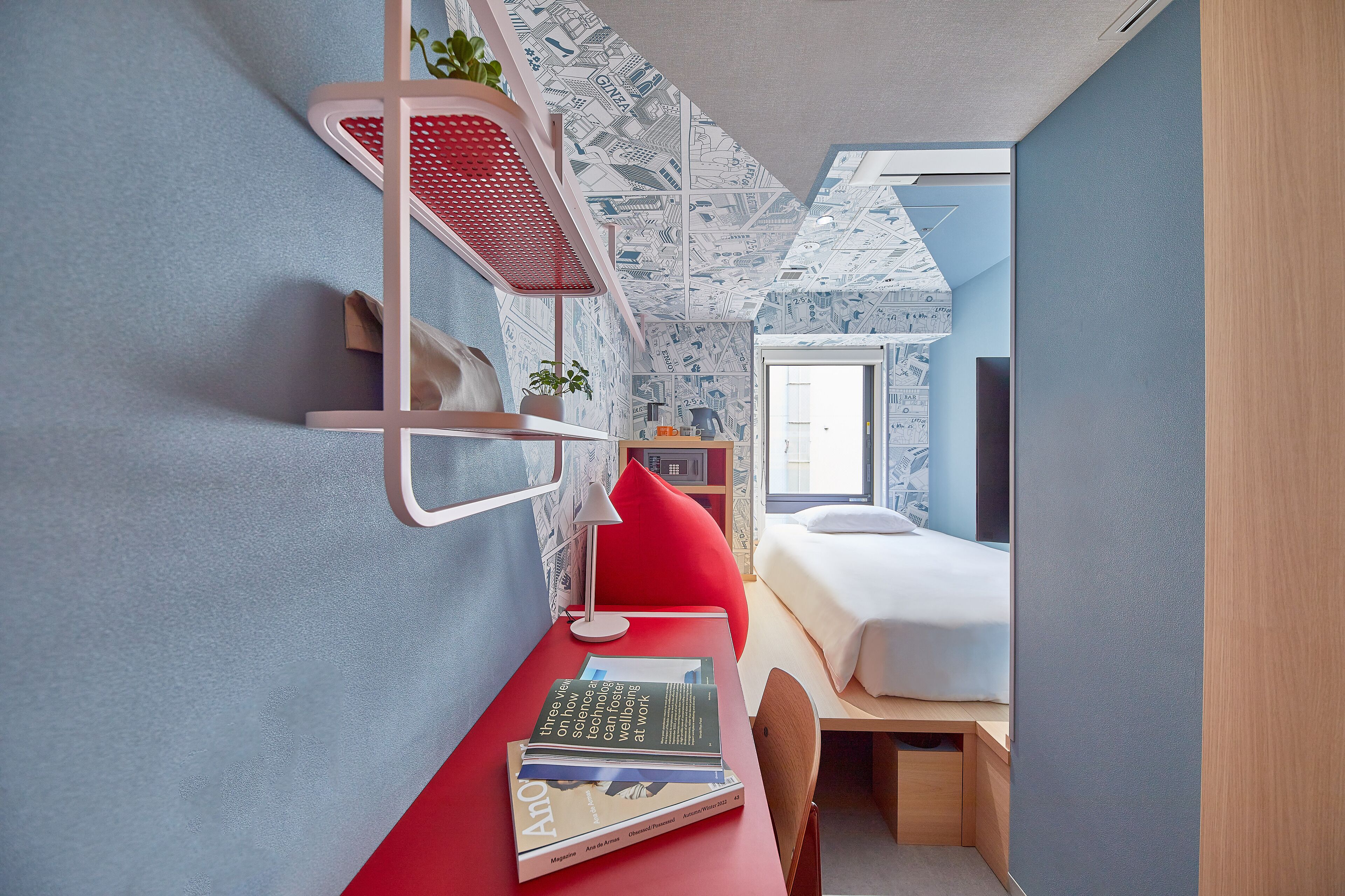 A compact room with a bed, a desk with books, a red chair, and a red shelf with a plant.