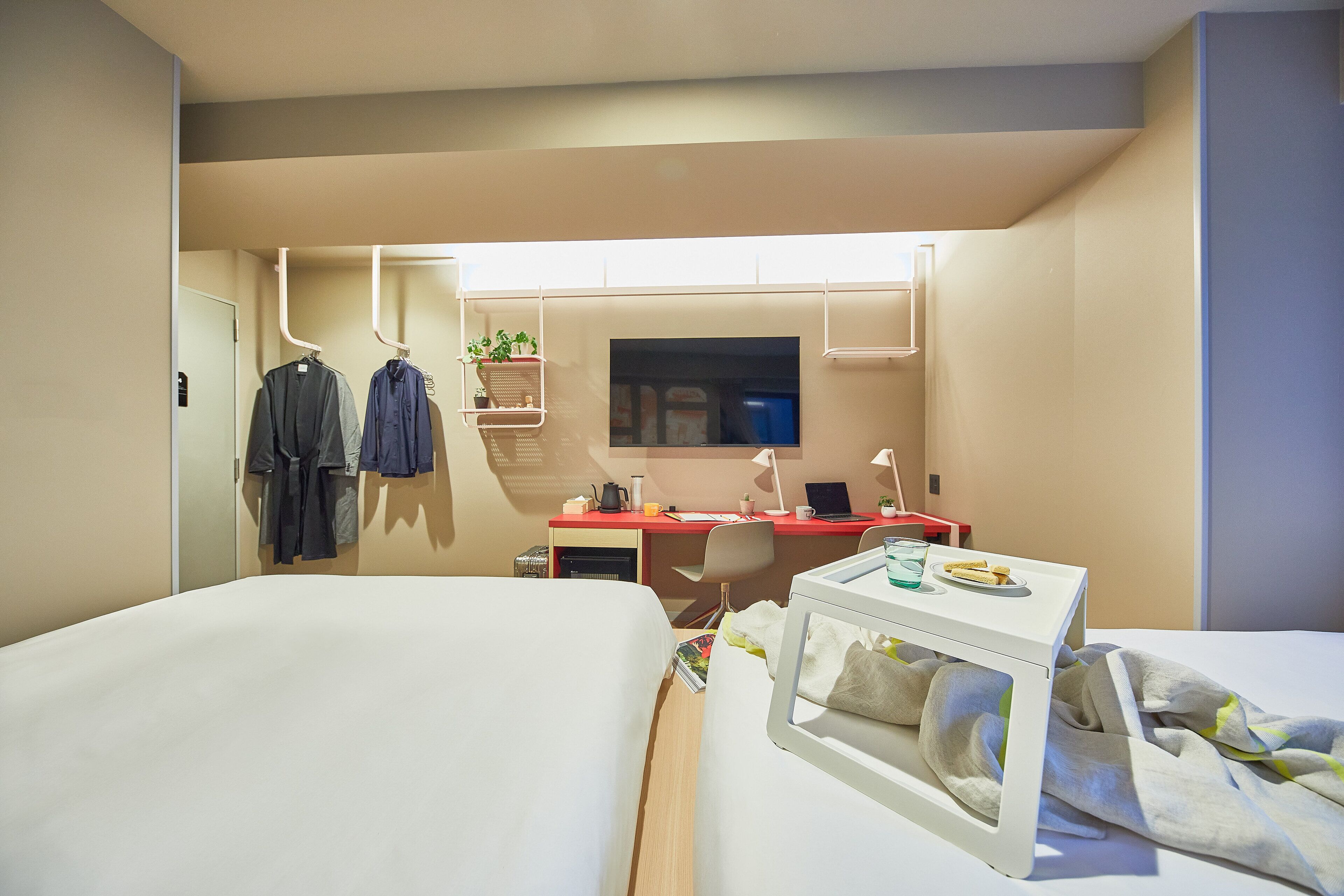 A compact hotel room with two beds, a desk with a computer, a small table with a glass of water, and a shelf with plants.