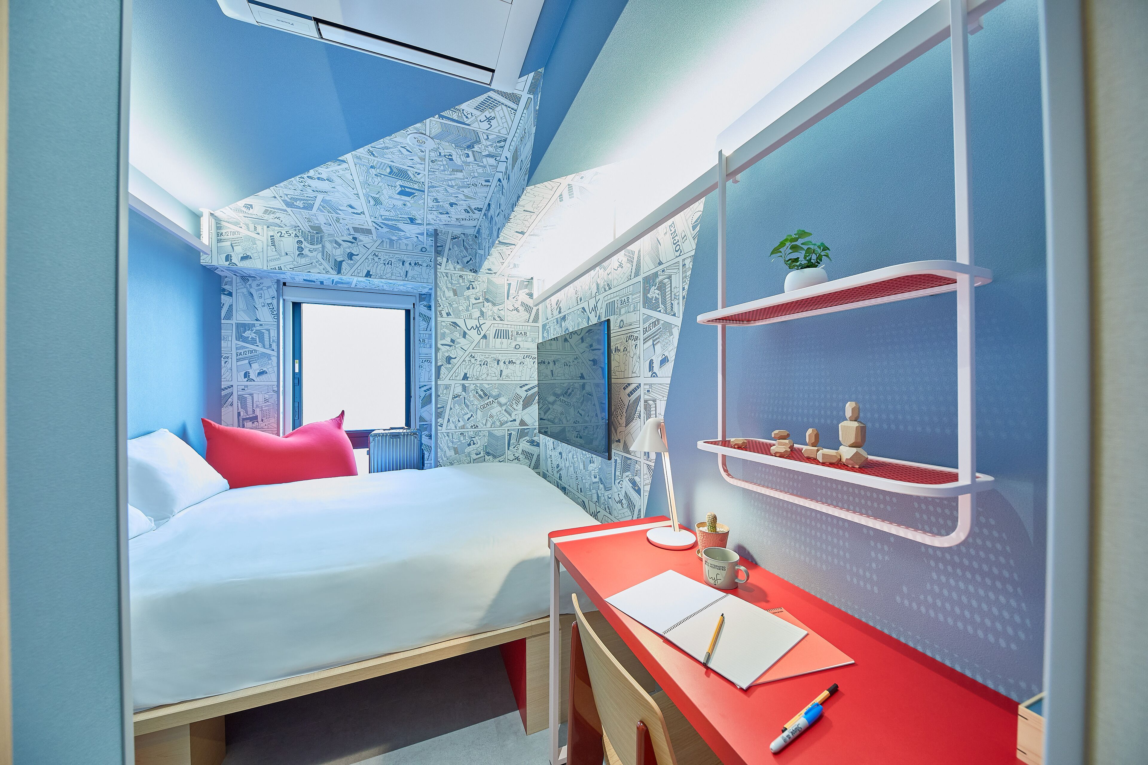 A bedroom with a bed, a desk, a chair, a window, and a wall with a blue and white pattern.