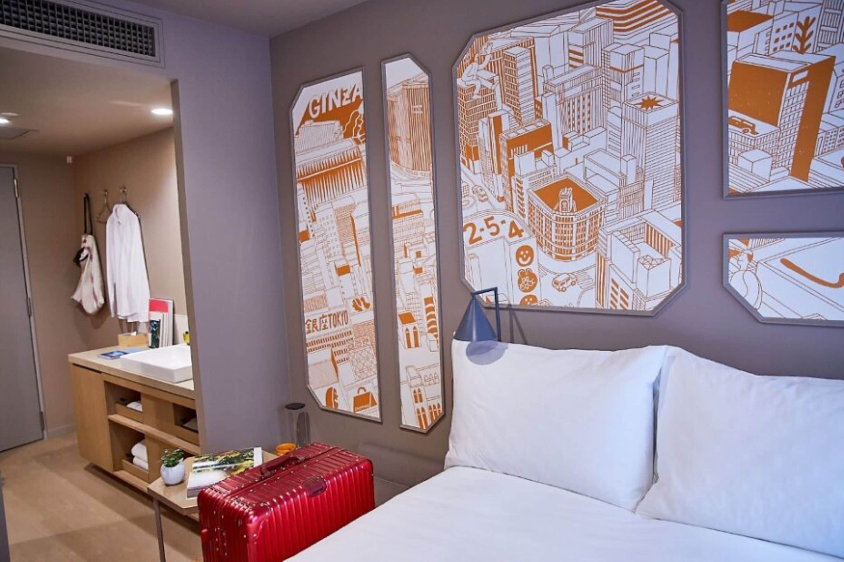 A hotel room with a bed, a red suitcase, a nightstand, and wall art depicting cityscapes.