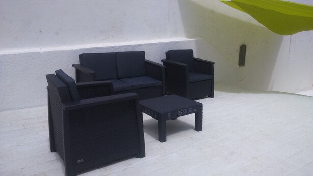 Una esquina de una habitación con una pared blanca y un tejido verde colgando. Hay un sofá negro y un sillón negro con una mesita delante.