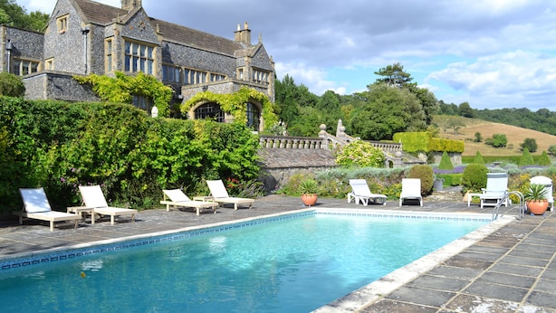 A stone building with a swimming pool, surrounded by greenery and a clear sky.