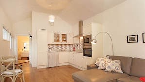 A modern kitchen with white cabinets, a microwave, and a stove. There is a dining area with a wooden table and chairs. A sofa with patterned cushions is in the living area.