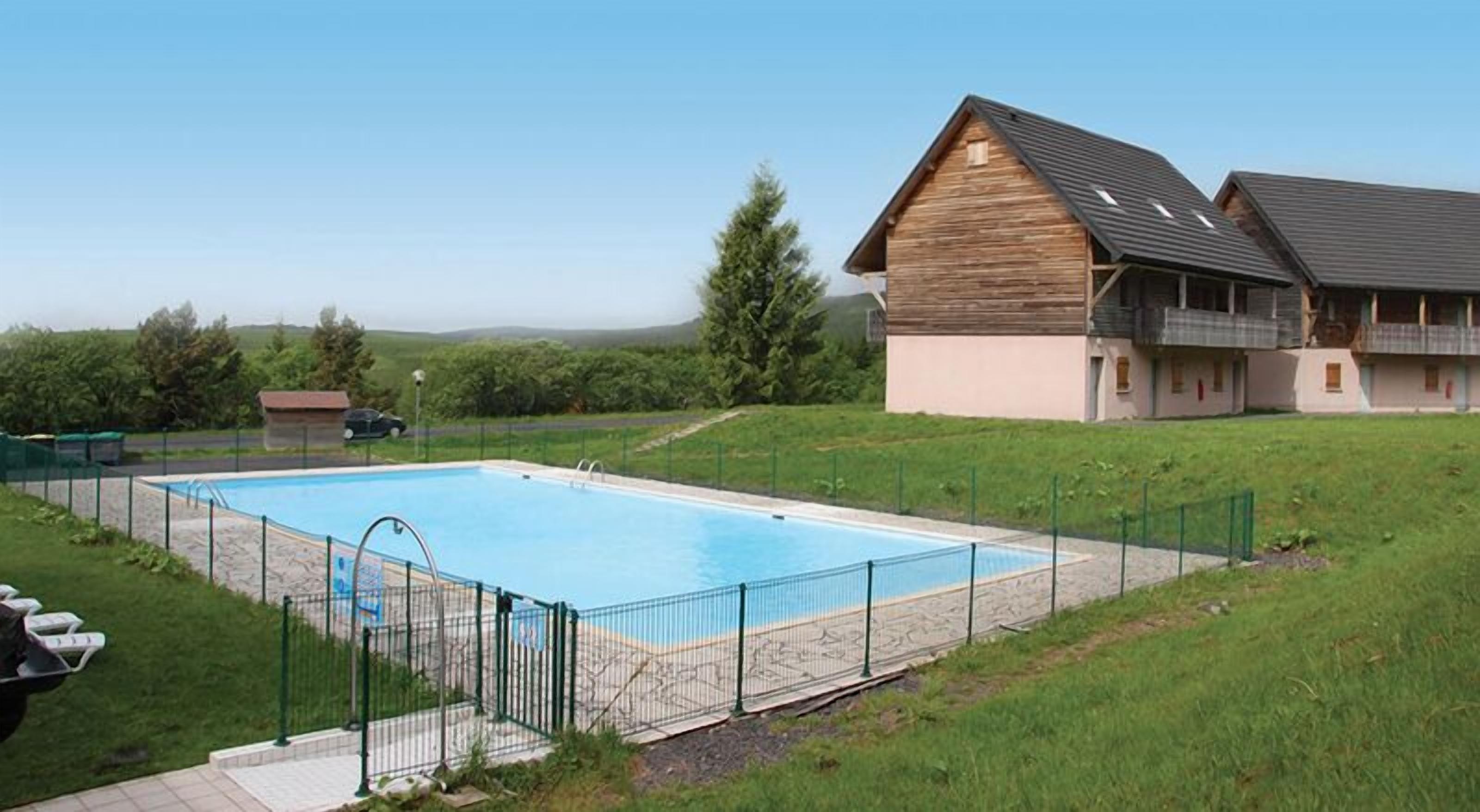 A rectangular swimming pool with a metal fence, surrounded by a grassy area and a building with a sloped roof.