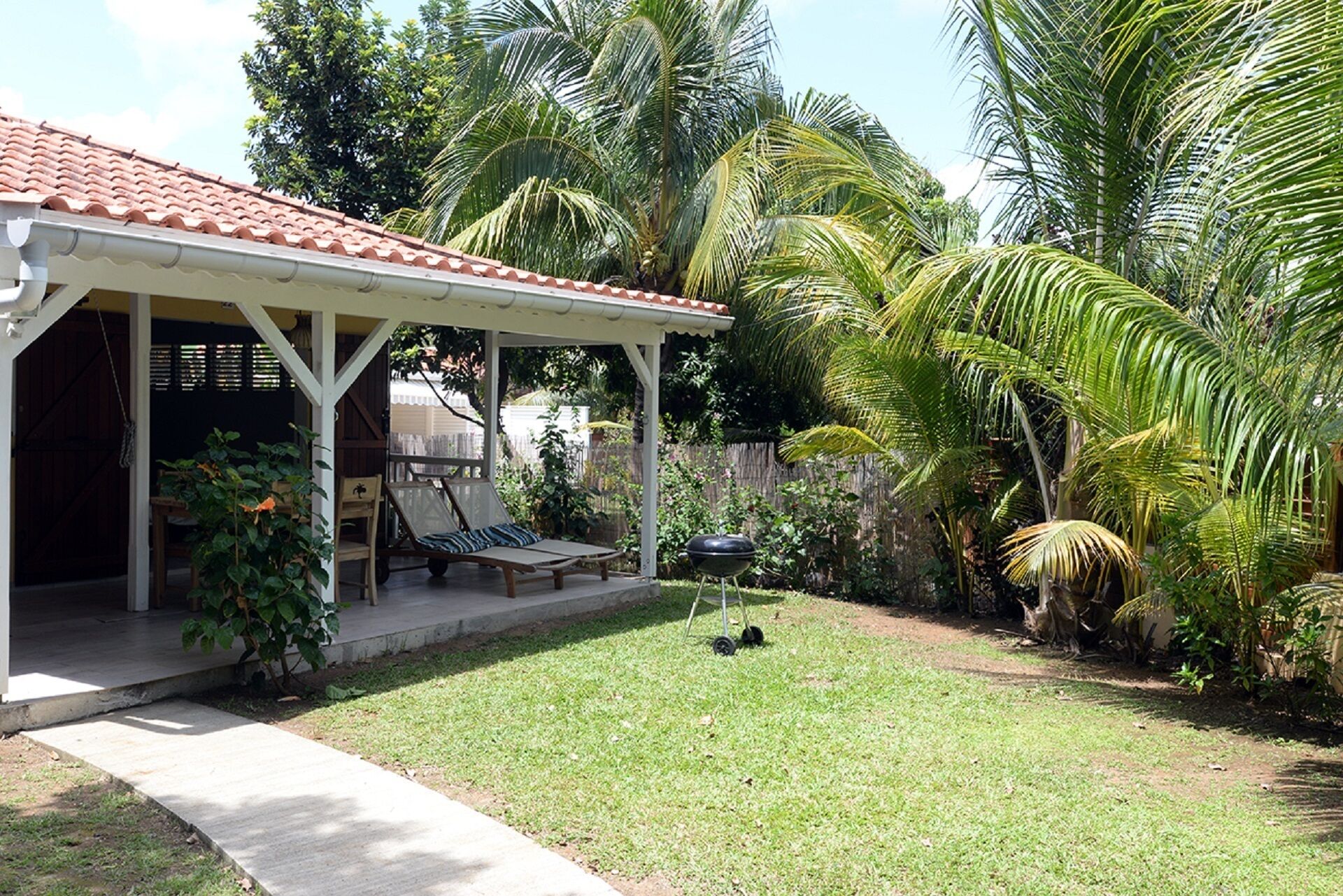 A covered patio with a grill, lounge chairs, and a small pool area surrounded by lush greenery and palm trees.