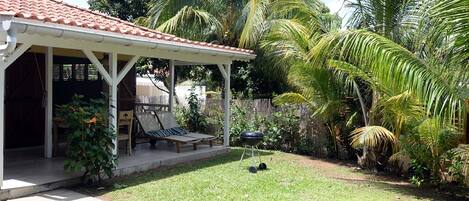 A covered patio with a grill, lounge chairs, and a small pool area surrounded by lush greenery and palm trees.