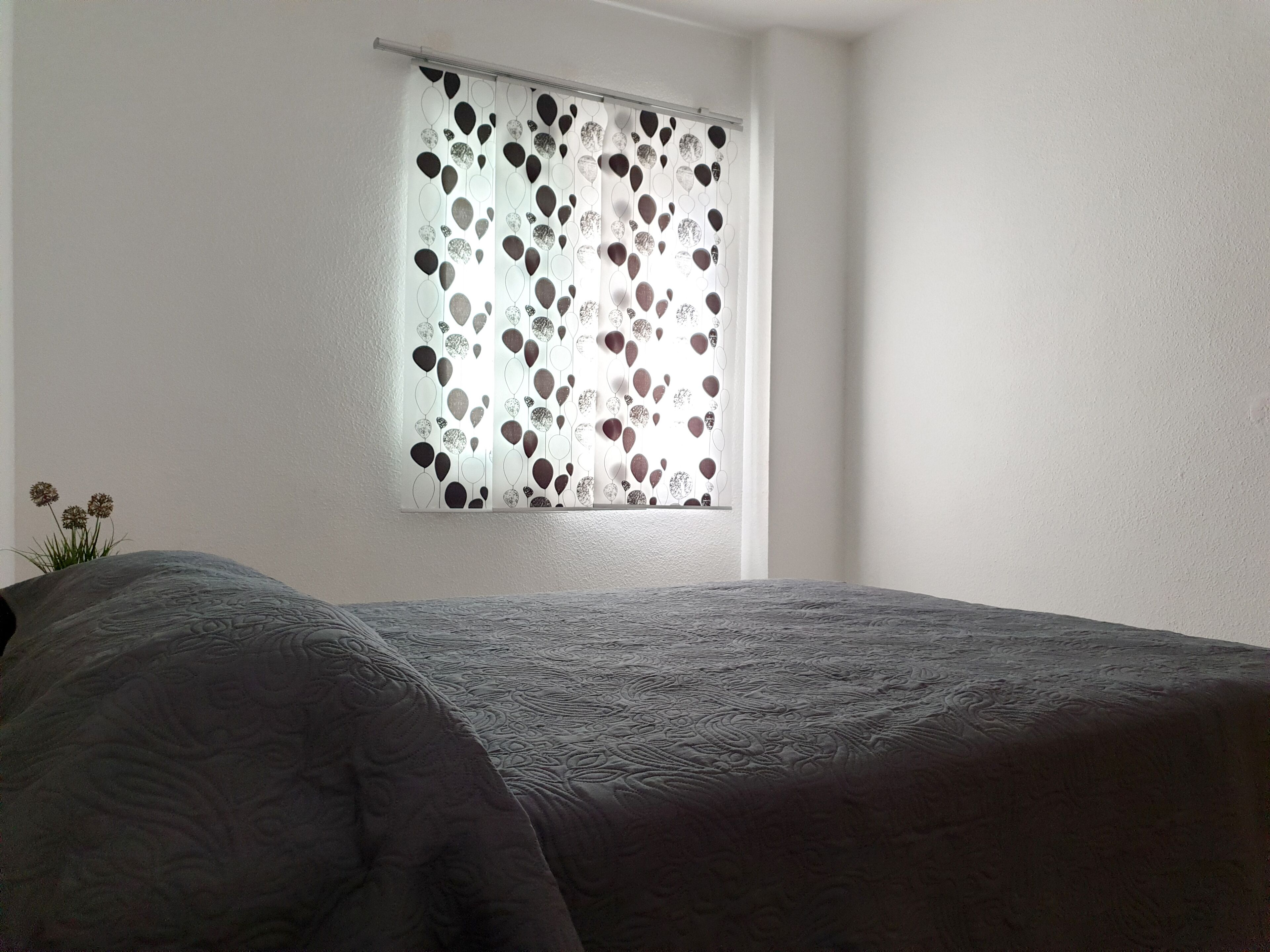 A bedroom with a bed, a window with decorative blinds, and a plant on the left.
