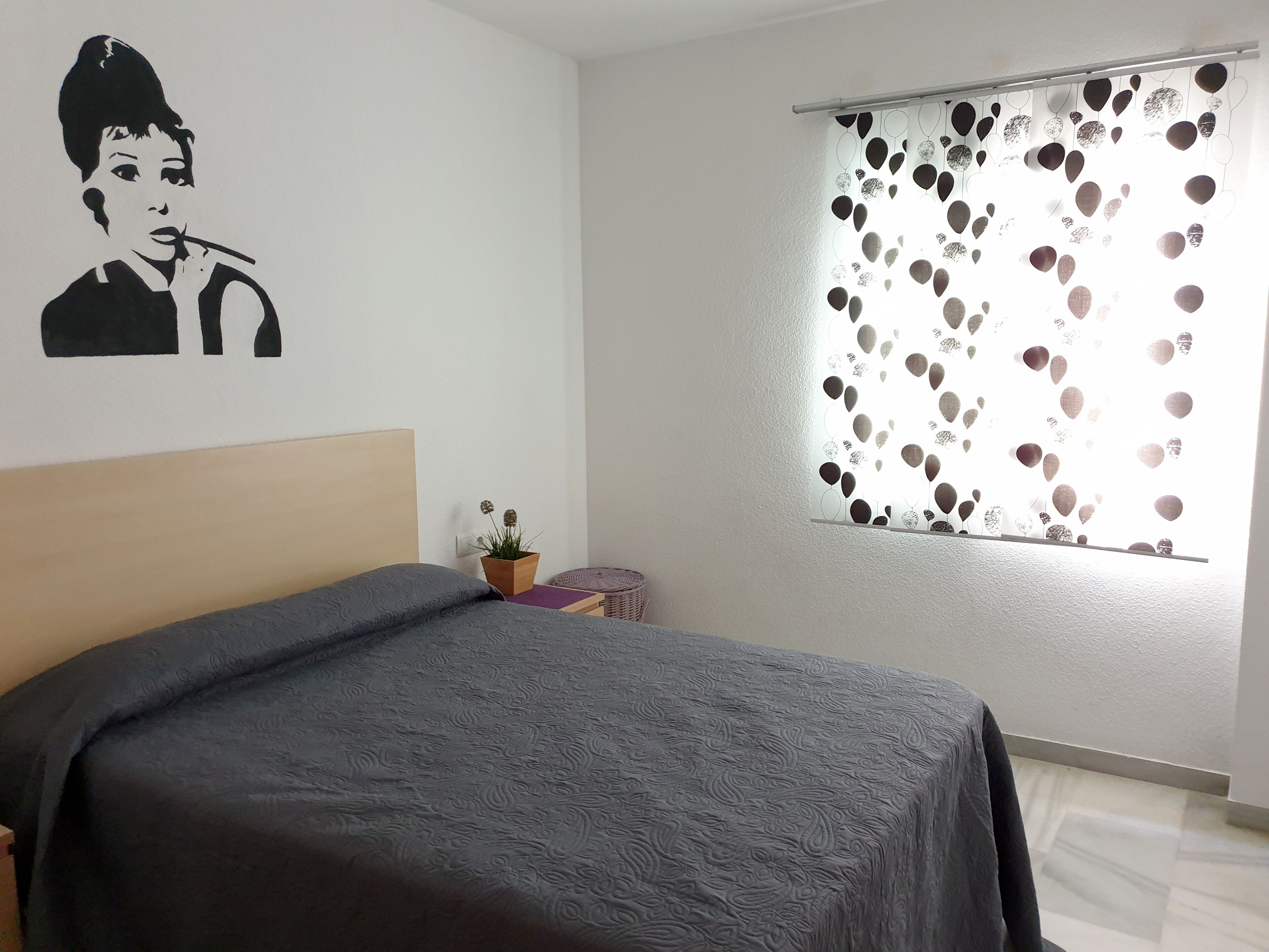 A bedroom with a bed, a wall decoration, a window with a patterned curtain, and a small potted plant.