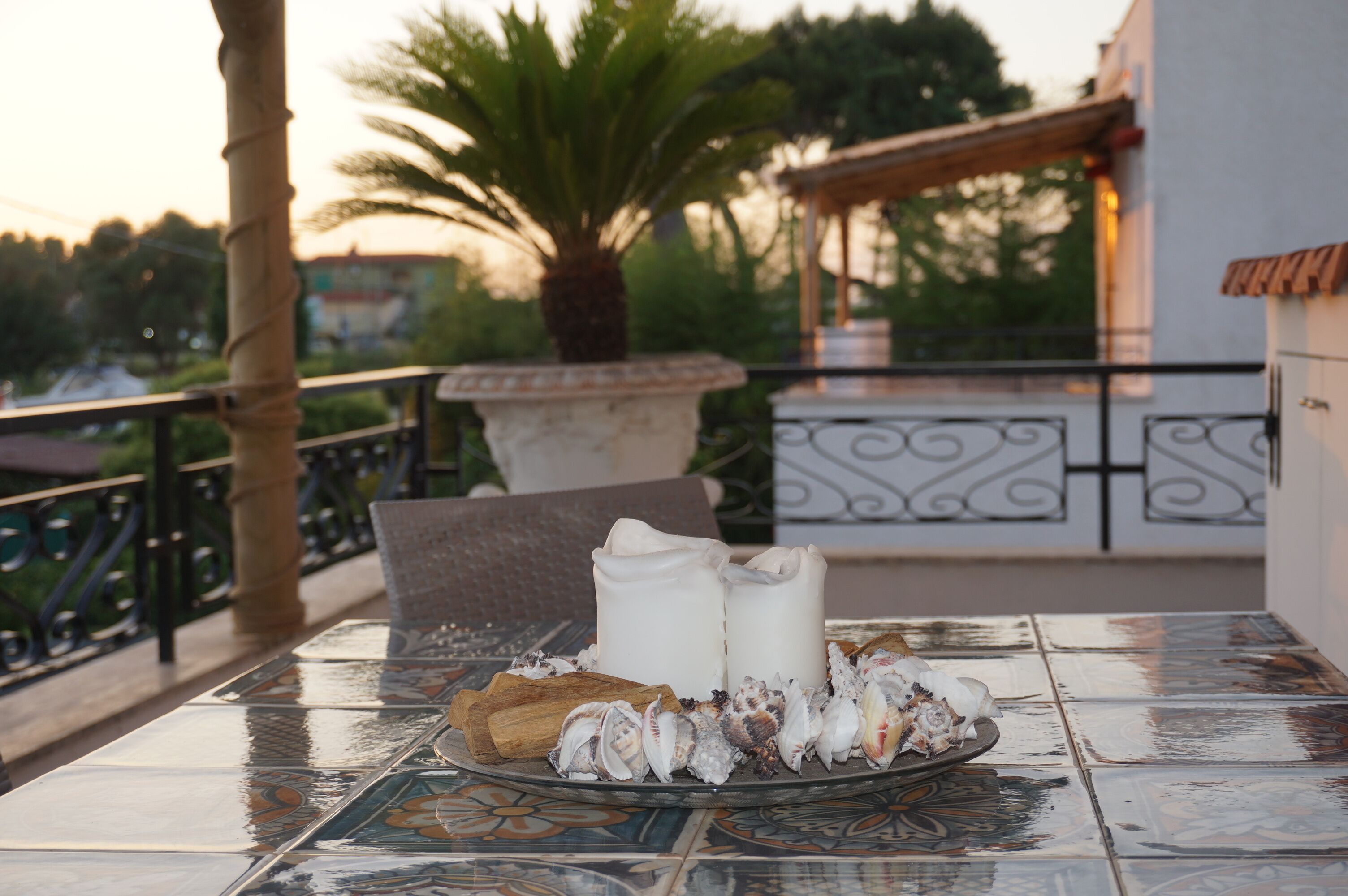 A table with a decorative tile pattern, a plate of seashells, and two candles on a balcony overlooking a palm tree and a building.