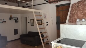 A loft-style room with a loft bed, a black sofa, a wooden ladder, a kitchen area with a stove, and a brick wall. - Loft in San Lorenzo, two minutes from the Acquario; optional parking (Genova)