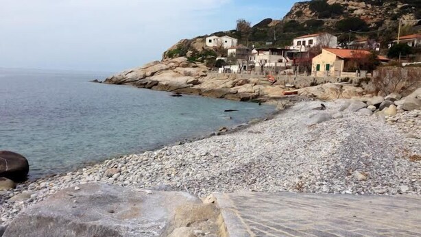 Une plage rocheuse avec une promenade dallée, des maisons sur une colline et une mer calme.