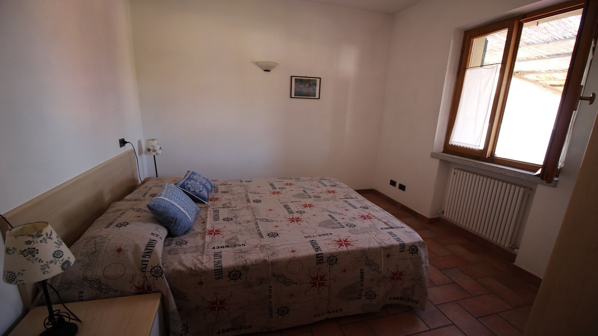 A small, single-bed room with a nautical-themed bedspread, a bedside lamp, and a window with a view of the outside.