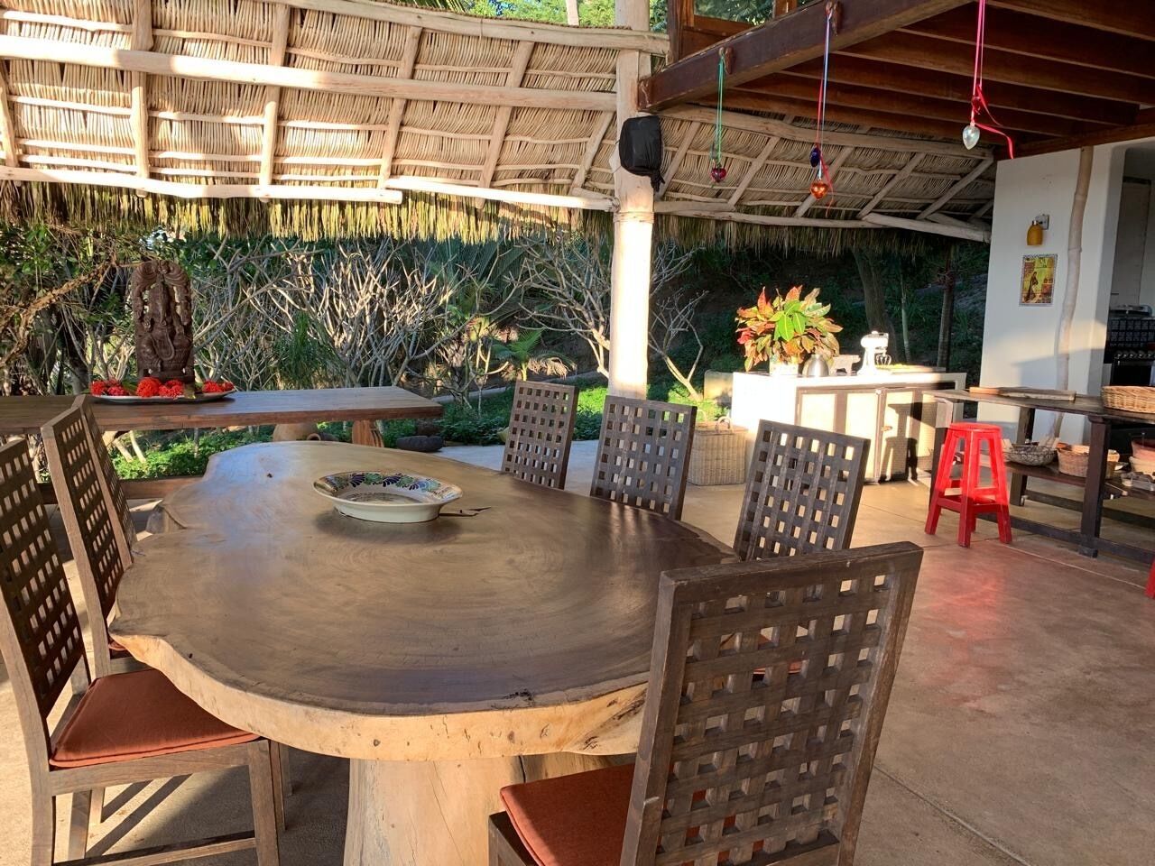 A round wooden table with six chairs, a thatched roof, and a garden with plants and a statue.