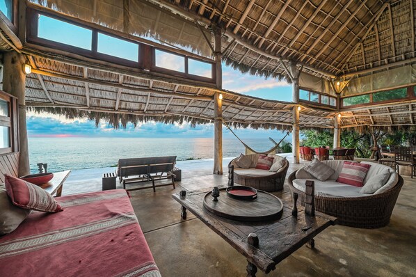 Un salón junto a la playa con techo de paja, muebles de mimbre y vistas al mar.