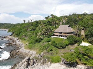 Une propriété située en bord de mer, avec une maison au toit de chaume, entourée d’une végétation luxuriante et de palmiers.
