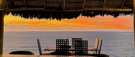 Un área de estar al aire libre con techo de paja, con vista a una masa de agua durante el atardecer.