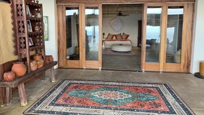 A spacious room with a large wooden bench, a patterned rug, and a view through glass doors to a balcony with outdoor furniture. - Gran Palapa Ocean Front Beach with Privacy Nature (SAYULITA)
