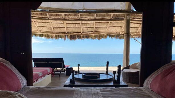 A bed with a view of the ocean, a thatched roof, and a small table with a tray.
