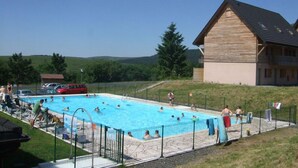 En swimmingpool med mennesker omkring, en rød bil og en træbygning i baggrunden.