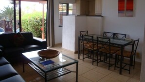Een woonkamer met een bank, salontafel en een eethoek met tafel en stoelen. Er is een schuifpui naar een balkon met planten.