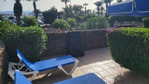 Een terras met blauwe loungestoelen, een stenen muur en uitzicht op palmbomen en geparkeerde auto's.