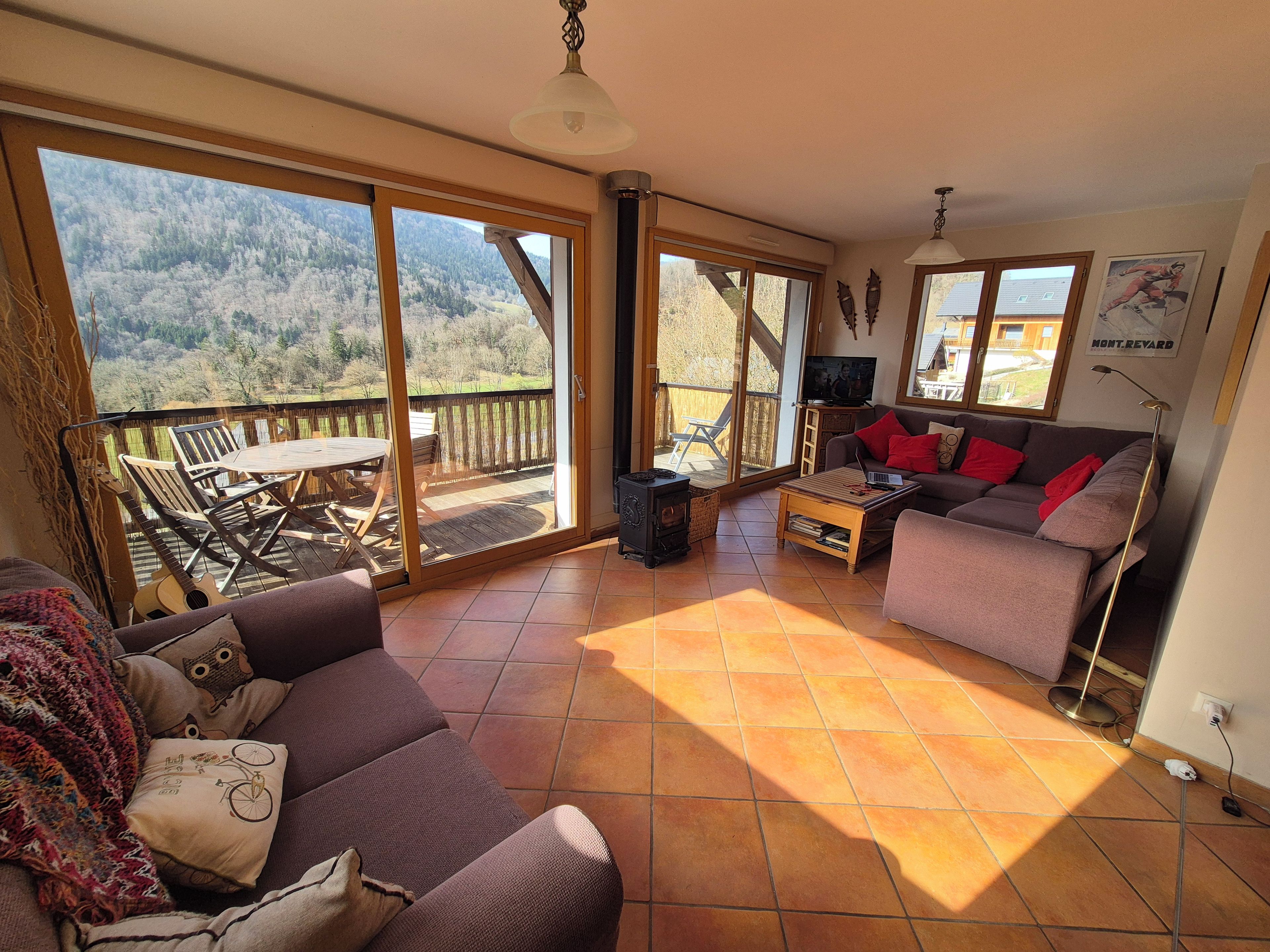 A living room with a sofa, a coffee table, and a TV. There is a balcony with a view of the mountains.