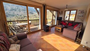 A living room with a sofa, a coffee table, and a TV. There is a balcony with a view of the mountains.