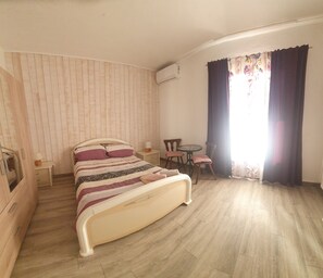 Quarto com cama, mesinha, duas cadeiras, criado-mudo e janela com cortinas.