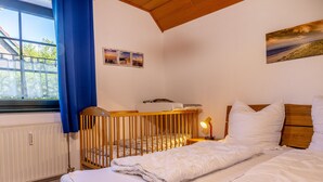 Ein Schlafzimmer mit einem Babybett, einem Bett mit weißer Bettwäsche, einem Nachttisch mit Lampe und einem Fenster mit blauen Vorhängen.