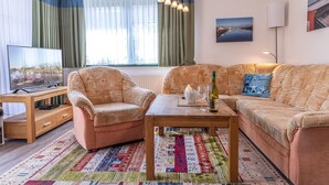 Ein Wohnzimmer mit einer Sofagarnitur, einem Couchtisch, einem Fernseher und einem großen Fenster mit Vorhängen.
