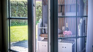 Blick durch eine Glastür in einen Raum mit einem gestreiften Sofa, einem kleinen Tisch mit einer Flasche und Blumen sowie einem Garten im Freien.