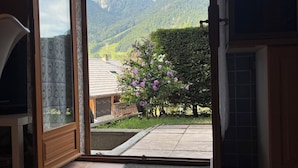 Una habitación con un amplio ventanal corredera que da a una vista de la montaña, un pequeño jardín con flores moradas y suelo de baldosas.