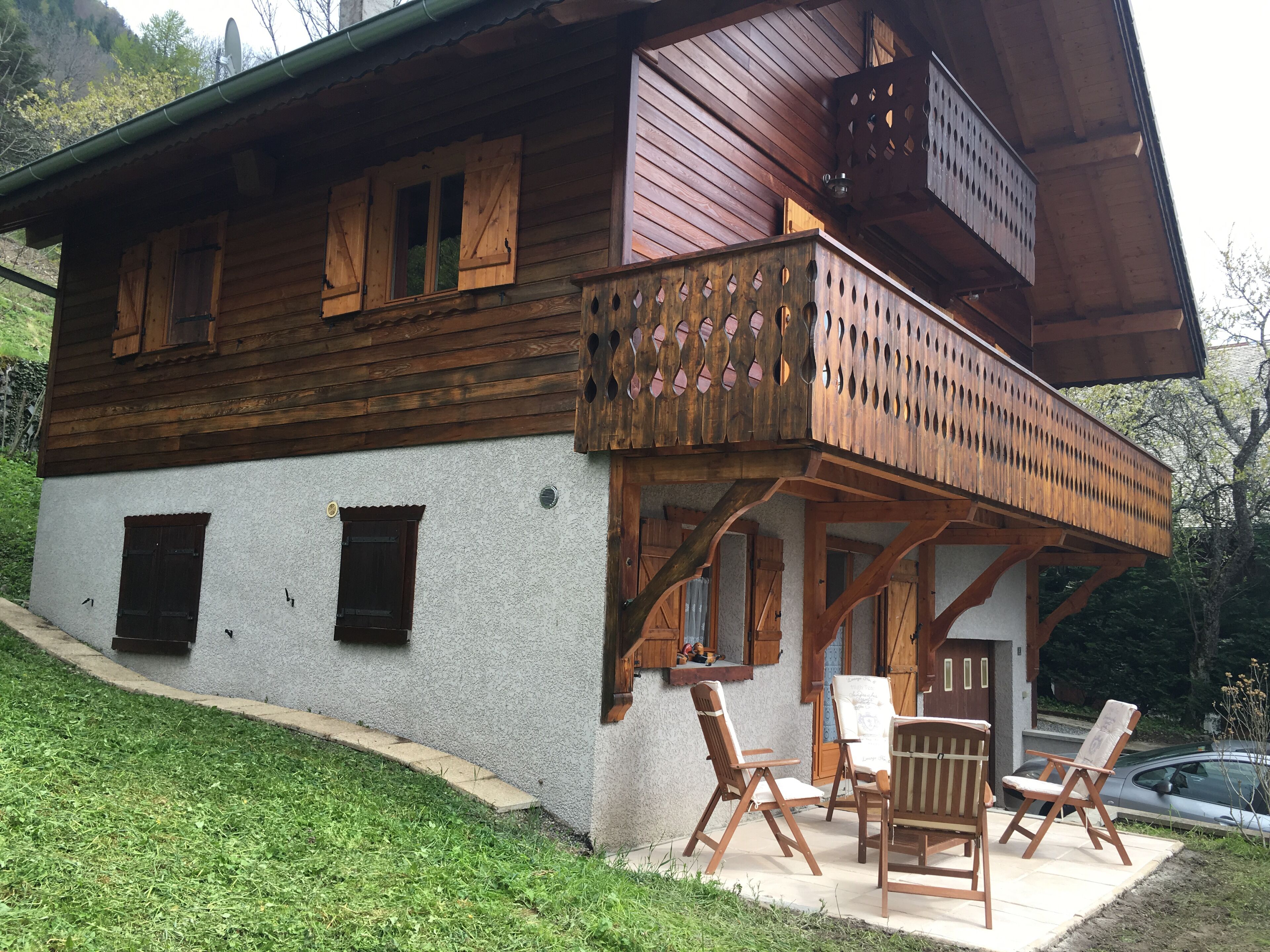 Una cabaña de madera con balcón, un patio con mobiliario exterior y un coche aparcado cerca.