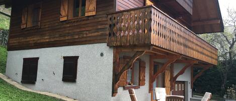 Una cabaña de madera con balcón, un patio con mobiliario exterior y un coche aparcado cerca.