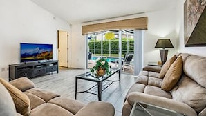 Un salón con sofá, una mesa de centro y televisor. Se ve una ventana con vista a una piscina y follaje.