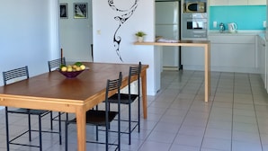 Een eetgedeelte met een houten tafel en stoelen, een schaal fruit en een wand met een zwart bloemenmotief.