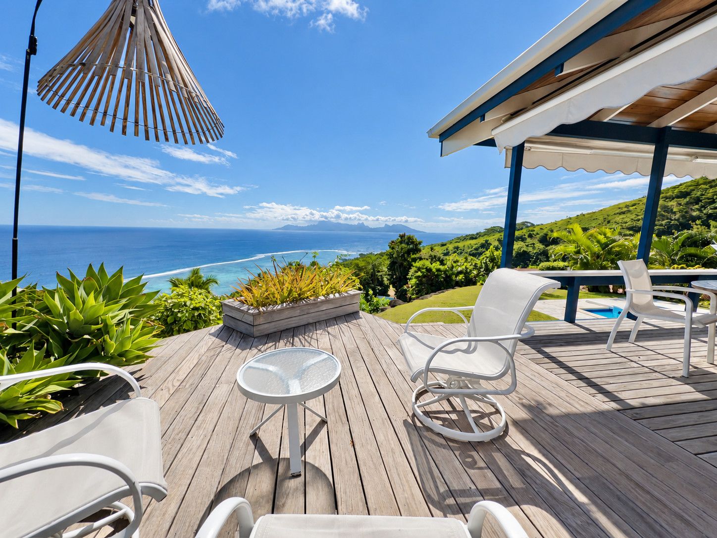 A wooden deck with outdoor furniture, a glass table, and a view of the ocean.