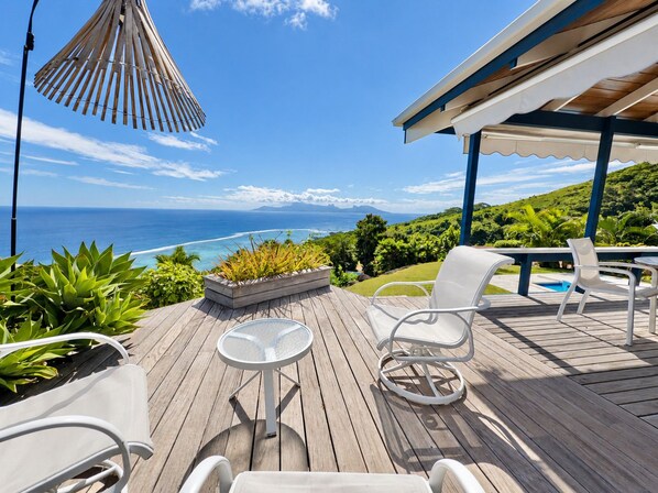 Una terraza de madera con mobiliario exterior, una mesa de vidrio y vista al océano.