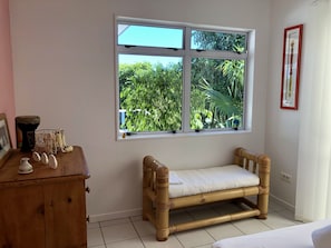 A room with a wooden bench, a small table with decorative items, and a window with a view of greenery. - Villa Tiare | Punaauia • Private Pool • Panoramic Moorea View • 4 Bedrooms (PUNAAUIA)
