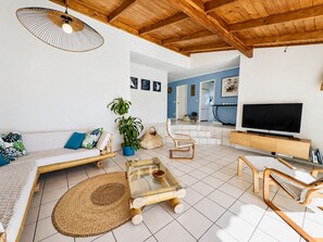 Una sala de estar moderna con techo de madera, un sofá con cojines, una mesa de centro, un televisor y un comedor con televisor.