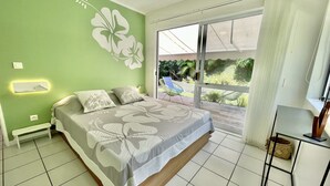 Een slaapkamer met een bed, een plafondventilator, een raam met uitzicht op groen en een tafeltje met een stoel.