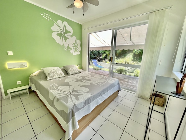 A bedroom with a bed, a ceiling fan, a window with a view of greenery, and a small table with a chair. - Villa Tiare | Punaauia • Private Pool • Panoramic Moorea View • 4 Bedrooms (PUNAAUIA)