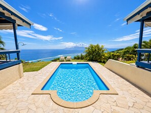 Una piscina rectangular con un diseño de mosaicos, rodeada de losas de piedra, y vista al océano y las montañas al fondo.
