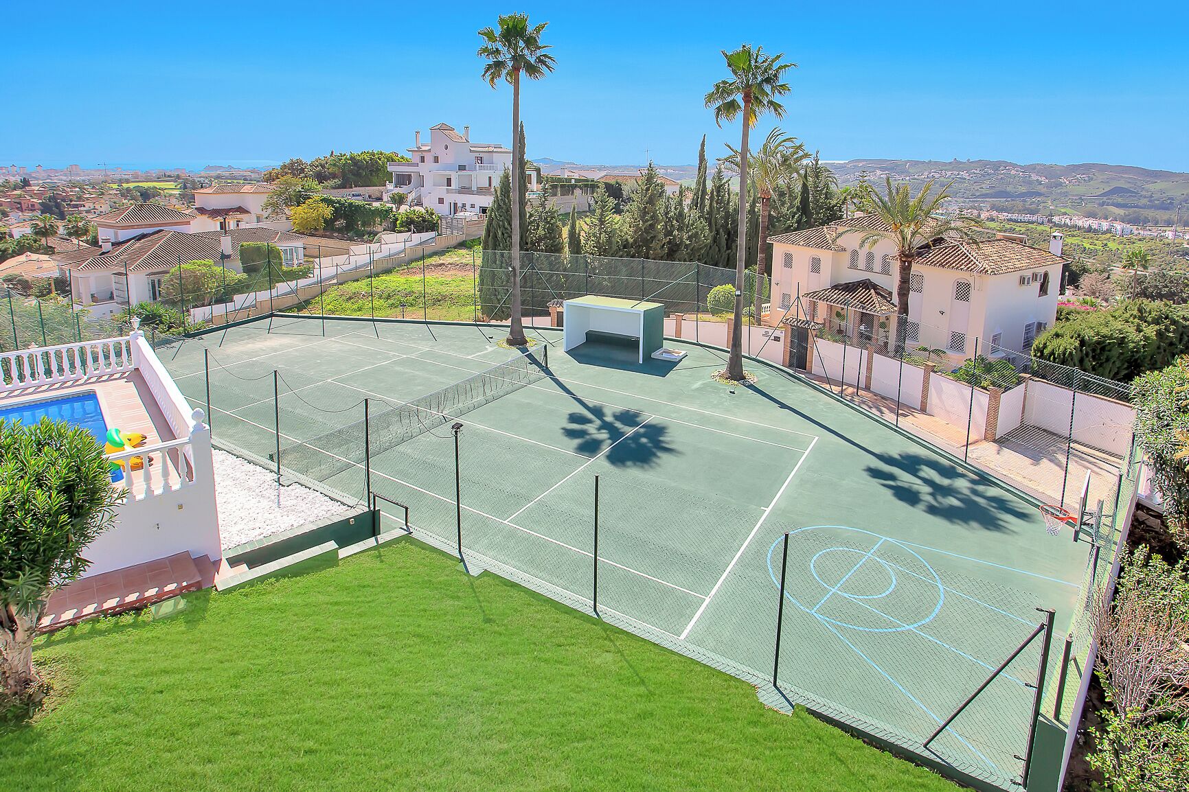 A tennis court with a net, surrounded by a fence, and a pool area with lounge chairs.