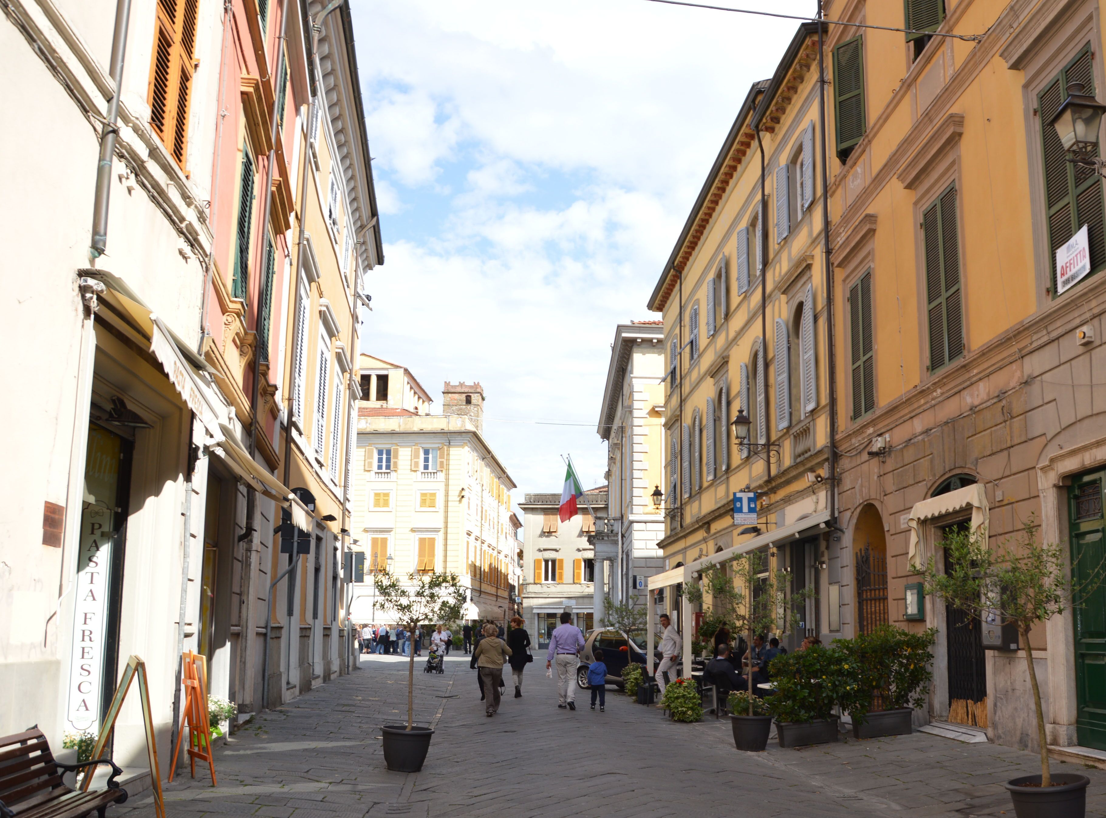 A narrow street with buildings on both sides, a flag, and people walking.