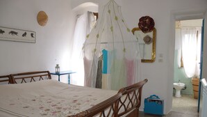 Una camera da letto con un letto in legno, un comodino, un armadio, uno specchio e un bagno parzialmente visibile attraverso una porta aperta, oltre a una poltrona appendibile.