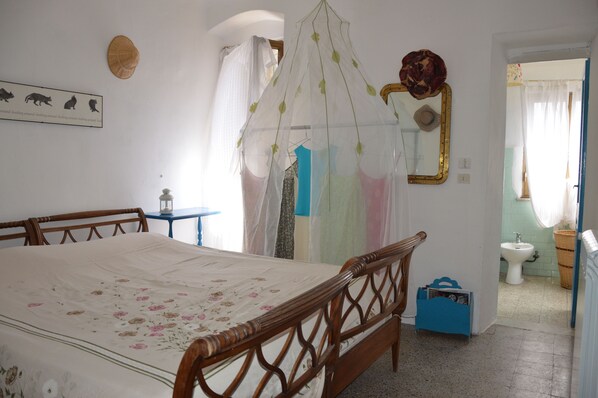 A bedroom with a wooden bed, a bedside table, a nightstand, a hanging chair, a wardrobe, a mirror, and a bathroom visible through an open door.