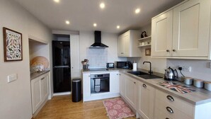 A compact kitchen with white cabinets, a black range hood, a microwave, a kettle, and a sink.