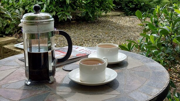 A round metal table with a coffee pot and two cups of coffee on it, set outdoors with a tree and bushes in the background.